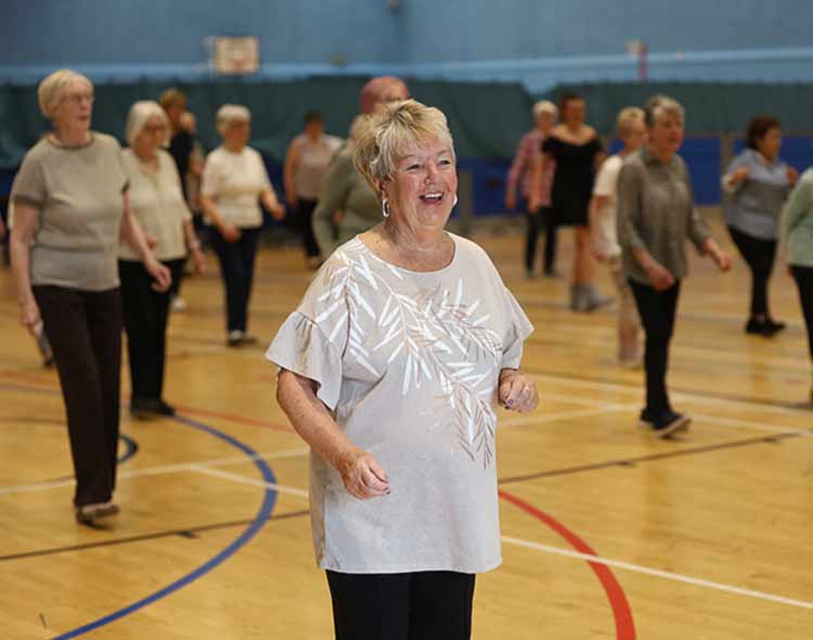 “A group of people taking part in a fitness or movement class inside a large indoor sports hall. Participants are spaced out across the gym floor, following an instructor and performing gentle exercise movements.”
