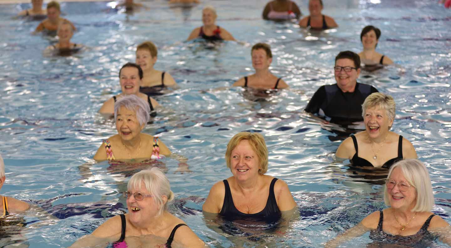 A group of people stand in waist‑deep water inside a swimming pool, participating in what appears to be a group aquatic exercise or water‑fitness class. They are spaced evenly across the pool, facing toward the front of the class. The water is clear and gently rippled around them. The background shows the edge of the pool, tiled flooring, and a poolside ladder.