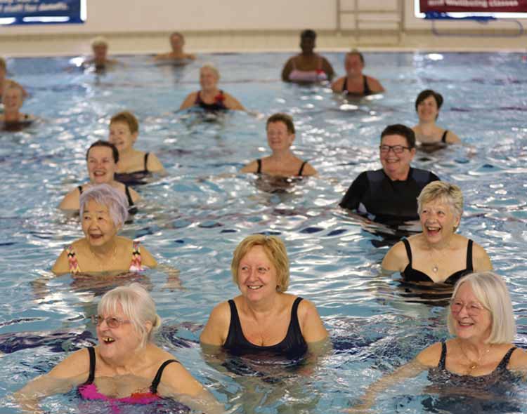 A group of people stand in waist‑deep water inside a swimming pool, participating in what appears to be a group aquatic exercise or water‑fitness class. They are spaced evenly across the pool, facing toward the front of the class. The water is clear and gently rippled around them. The background shows the edge of the pool, tiled flooring, and a poolside ladder.