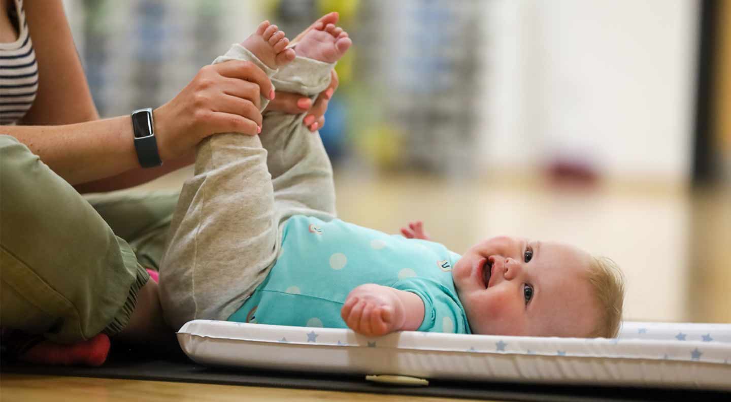 An adult sits on the floor and gently holds a baby’s legs, lifting them as the baby lies on a padded mat. The baby is wearing a turquoise top with light-coloured trousers and is looking upward. The scene is in a exercise setting.