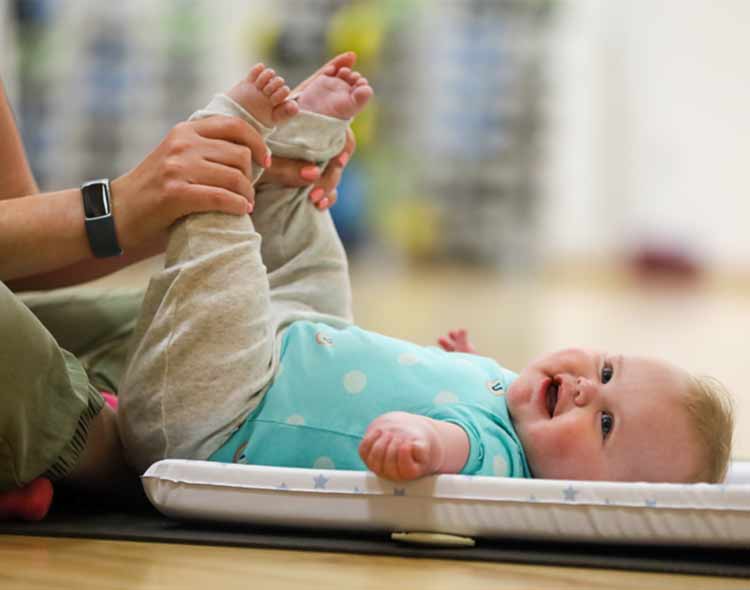 An adult sits on the floor and gently holds a baby’s legs, lifting them as the baby lies on a padded mat. The baby is wearing a turquoise top with light-coloured trousers and is looking upward. The scene appears to take place in a exercise setting.