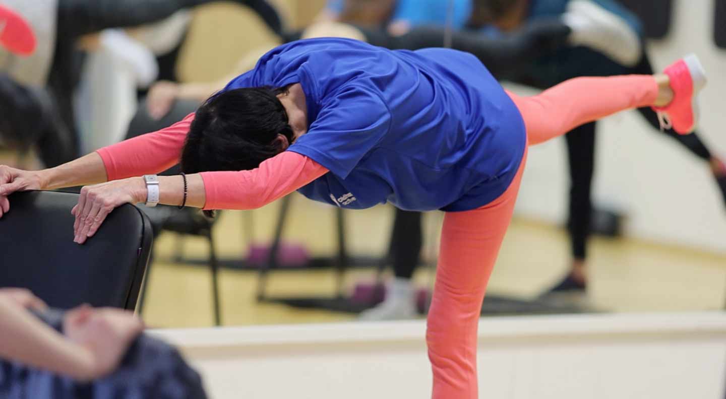 A participant in a chair‑assisted exercise class leaning forward with outstretched arms while lifting one leg straight behind on a balance pose.