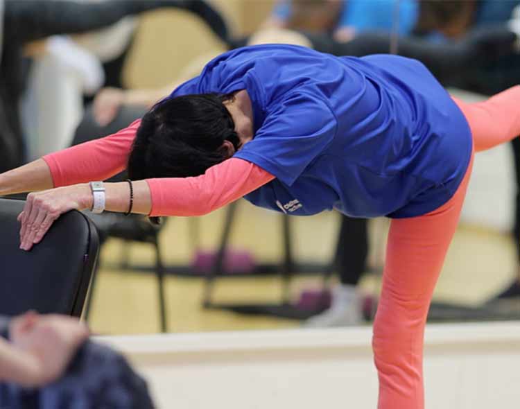 A participant in a chair‑assisted exercise class leaning forward with outstretched arms while lifting one leg straight behind on a balance pose.