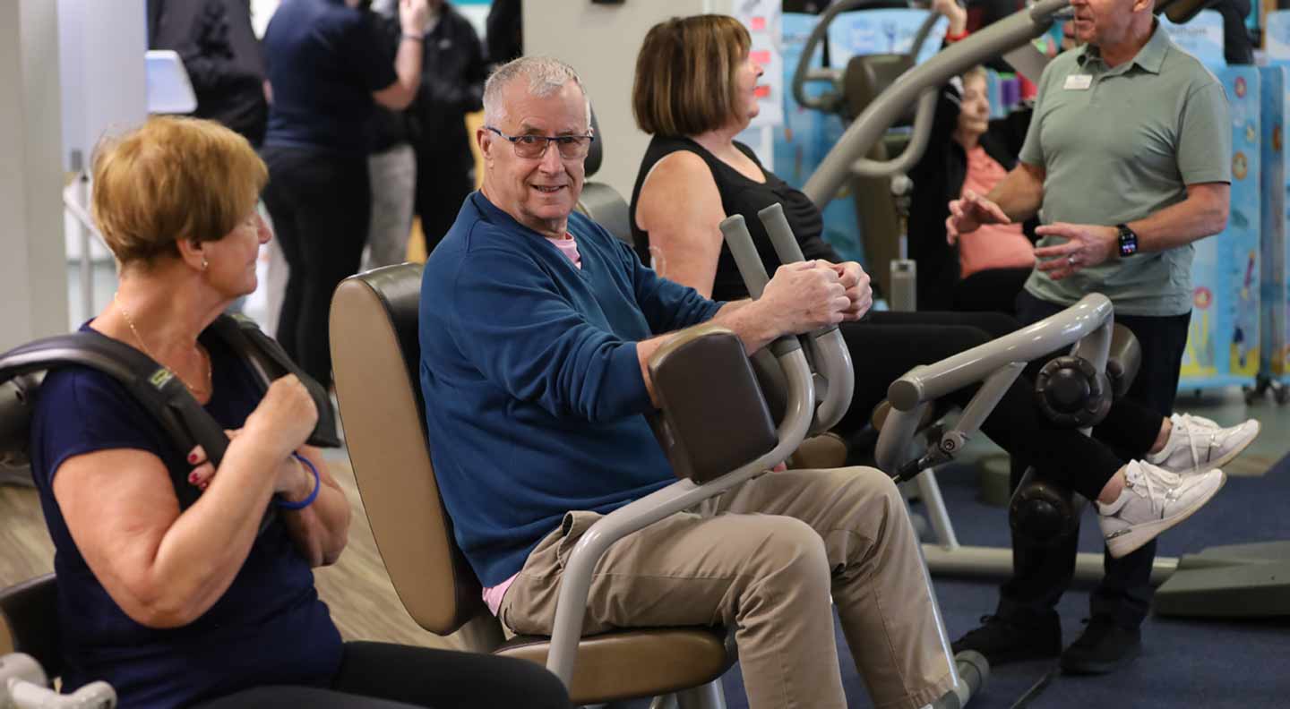 A group of individuals is seated on various exercise machines inside a gym or fitness center. In the foreground, one person uses a shoulder‑press–style machine, while another person sits on a seated rowing machine holding the handles. A third person sits on a similar machine nearby. In the background, additional people stand and talk near more exercise equipment. The setting is bright and busy, with multiple workout machines, blue carpeting, and informational posters visible throughout the space.