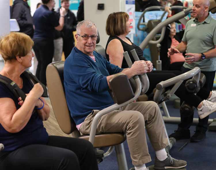 A group of individuals is seated on various exercise machines inside a gym or fitness center. In the foreground, one person uses a shoulder‑press–style machine, while another person sits on a seated rowing machine holding the handles. A third person sits on a similar machine nearby. In the background, additional people stand and talk near more exercise equipment. The setting is bright and busy, with multiple workout machines, blue carpeting, and informational posters visible throughout the space.