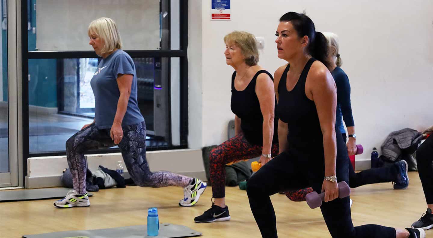 A group fitness class performing forward lunges while holding dumbbells in a studio with exercise mats and equipment on the floor.