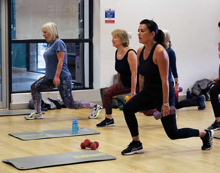 A group fitness class performing forward lunges while holding dumbbells in a studio with exercise mats and equipment on the floor.
