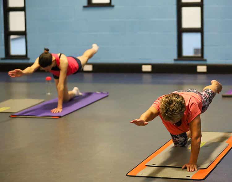 Two people in a fitness studio are practicing a balance exercise on mats. Each person is positioned on hands and knees, extending one arm forward and the opposite leg backward. The studio has a smooth floor, blue walls, and several small windows. The individuals are spaced apart on different colored mats, with water bottles and other small items nearby.