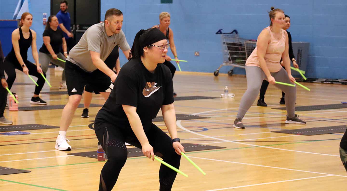 A group fitness class taking place in an indoor sports hall. Participants are positioned on exercise mats and holding bright green drumsticks, performing a squat movement as part of a Pound-style workout.
