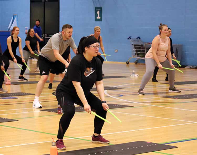 A group fitness class taking place in an indoor sports hall. Participants are positioned on exercise mats and holding bright green drumsticks, performing a squat movement as part of a Pound-style workout.