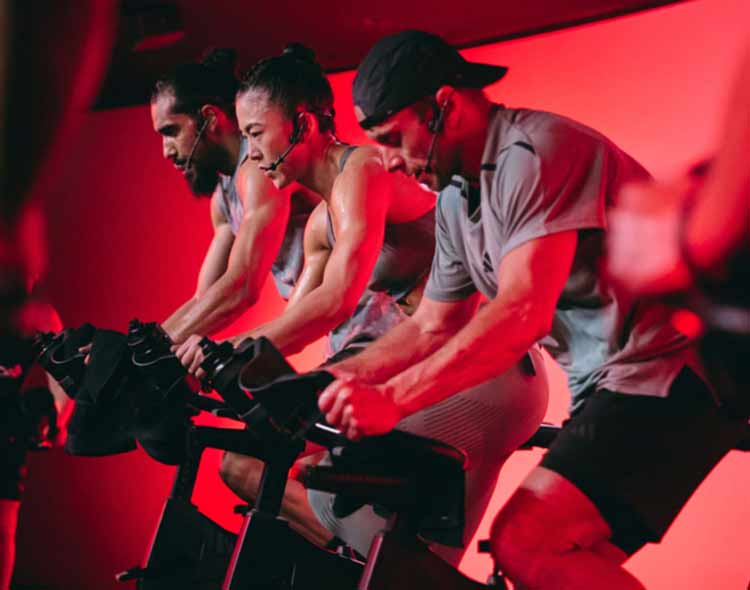 Several people ride stationary bikes in a dimly lit indoor cycling studio with a strong red light illuminating the room. The group is leaned forward in an intense riding position, gripping the handlebars as they pedal. The bikes are arranged closely together, creating a focused, energetic workout atmosphere.