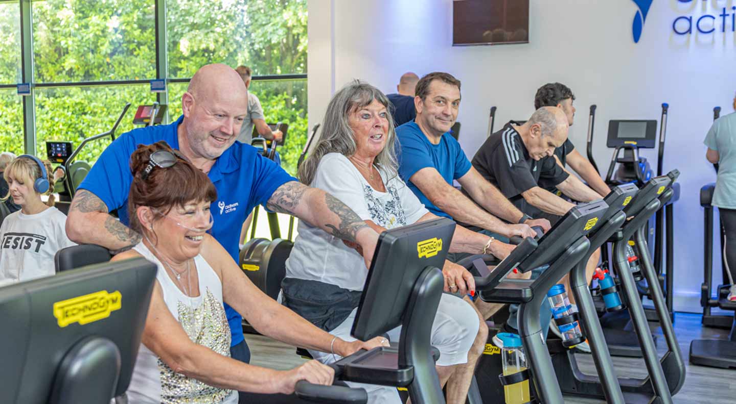 A group of people use stationary exercise bikes inside a bright, modern fitness studio. Several individuals are positioned in a row, each holding the handlebars while cycling. A person wearing a blue top appears to be providing guidance or support to one of the participants. Large windows on the left side reveal green trees outside, allowing natural light to fill the room. The interior features light-colored walls, fitness branding on the right, and a row of additional equipment in the background. The scene captures an active, social workout environment.
