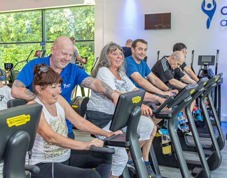 A group of people use stationary exercise bikes inside a bright, modern fitness studio. Several individuals are positioned in a row, each holding the handlebars while cycling. A person wearing a blue top appears to be providing guidance or support to one of the participants. Large windows on the left side reveal green trees outside, allowing natural light to fill the room. The interior features light-colored walls, fitness branding on the right, and a row of additional equipment in the background. The scene captures an active, social workout environment.