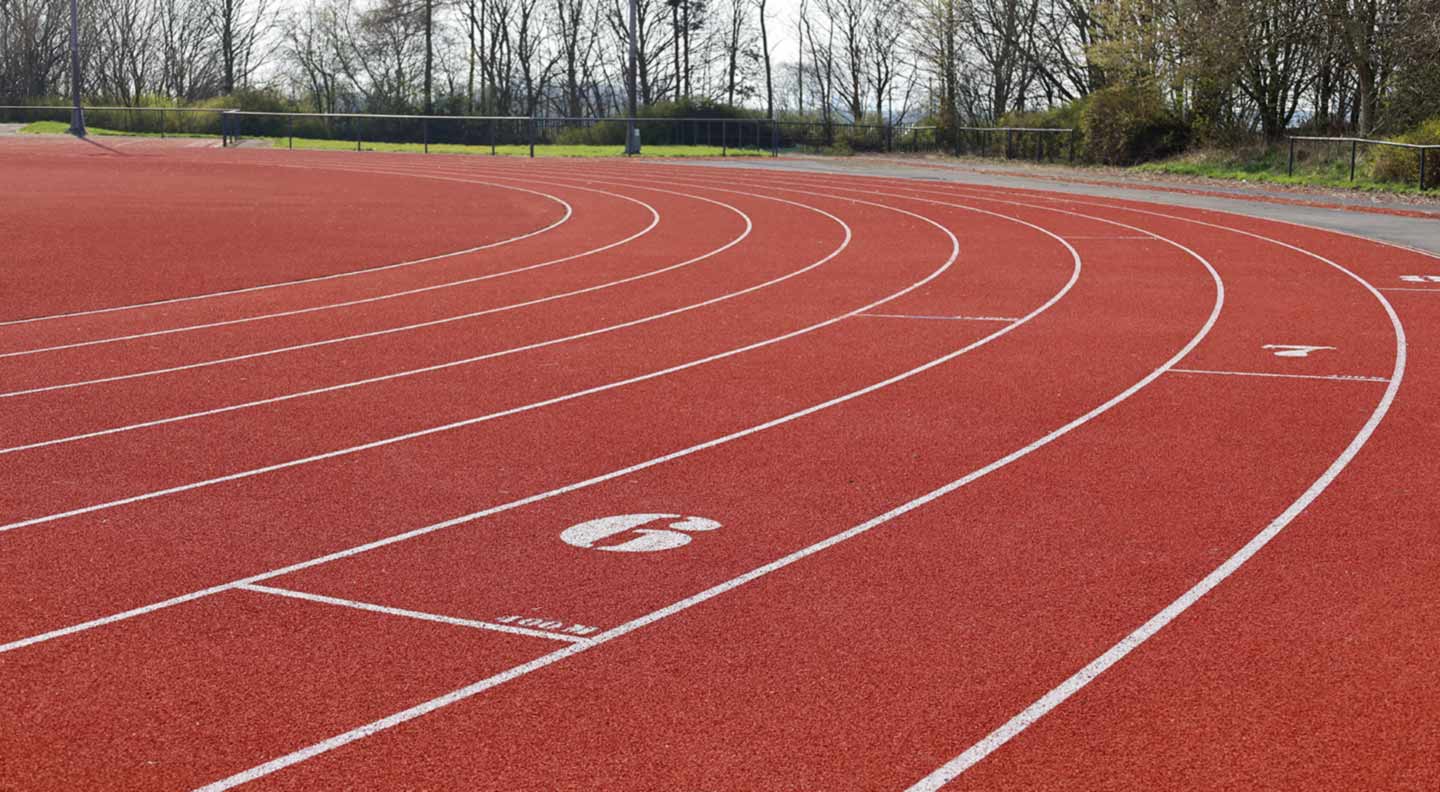 A red outdoor running track with marked lanes curving to the left. The surface shows clear white lane numbers and lines, and trees border the track in the background.”