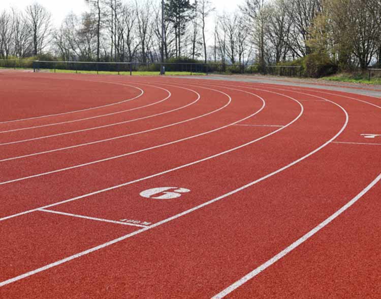 A red outdoor running track with marked lanes curving to the left. The surface shows clear white lane numbers and lines, and trees border the track in the background.”