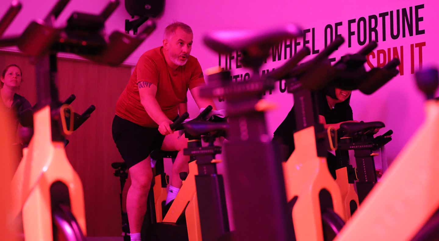 Several people take part in an indoor cycling class in a room lit with bright pink and purple lighting. Stationary bikes are arranged in rows, and one participant near the center is leaning forward while pedalling on a yellow-and-black bike. Text on the wall reads “LIFE IS A WHEEL OF FORTUNE… SPIN IT.” Additional bikes and participants are visible in the background, creating a busy, energetic studio atmosphere.
