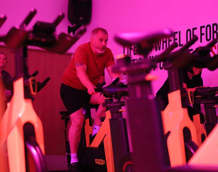 Several people take part in an indoor cycling class in a room lit with bright pink and purple lighting. Stationary bikes are arranged in rows, and one participant near the center is leaning forward while pedalling on a yellow-and-black bike. Text on the wall reads “LIFE IS A WHEEL OF FORTUNE… SPIN IT.” Additional bikes and participants are visible in the background, creating a busy, energetic studio atmosphere.