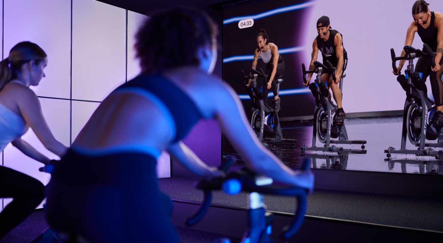 People participate in an indoor cycling class in a dark studio illuminated with soft purple lighting. Several individuals ride stationary bikes facing a large screen at the front of the room, which displays three instructors also riding bikes. The setup creates an immersive, high‑tech fitness environment with reflections visible on the glossy floor.