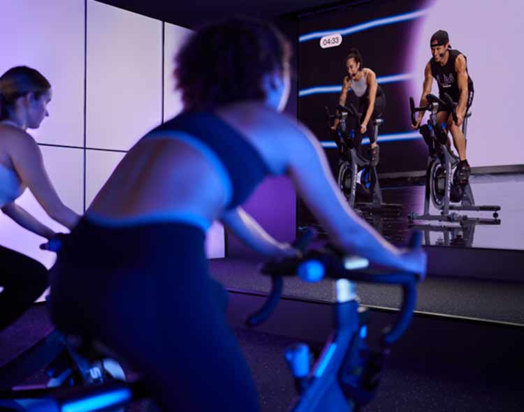 People participate in an indoor cycling class in a dark studio illuminated with soft purple lighting. Several individuals ride stationary bikes facing a large screen at the front of the room, which displays three instructors also riding bikes. The setup creates an immersive, high‑tech fitness environment with reflections visible on the glossy floor.