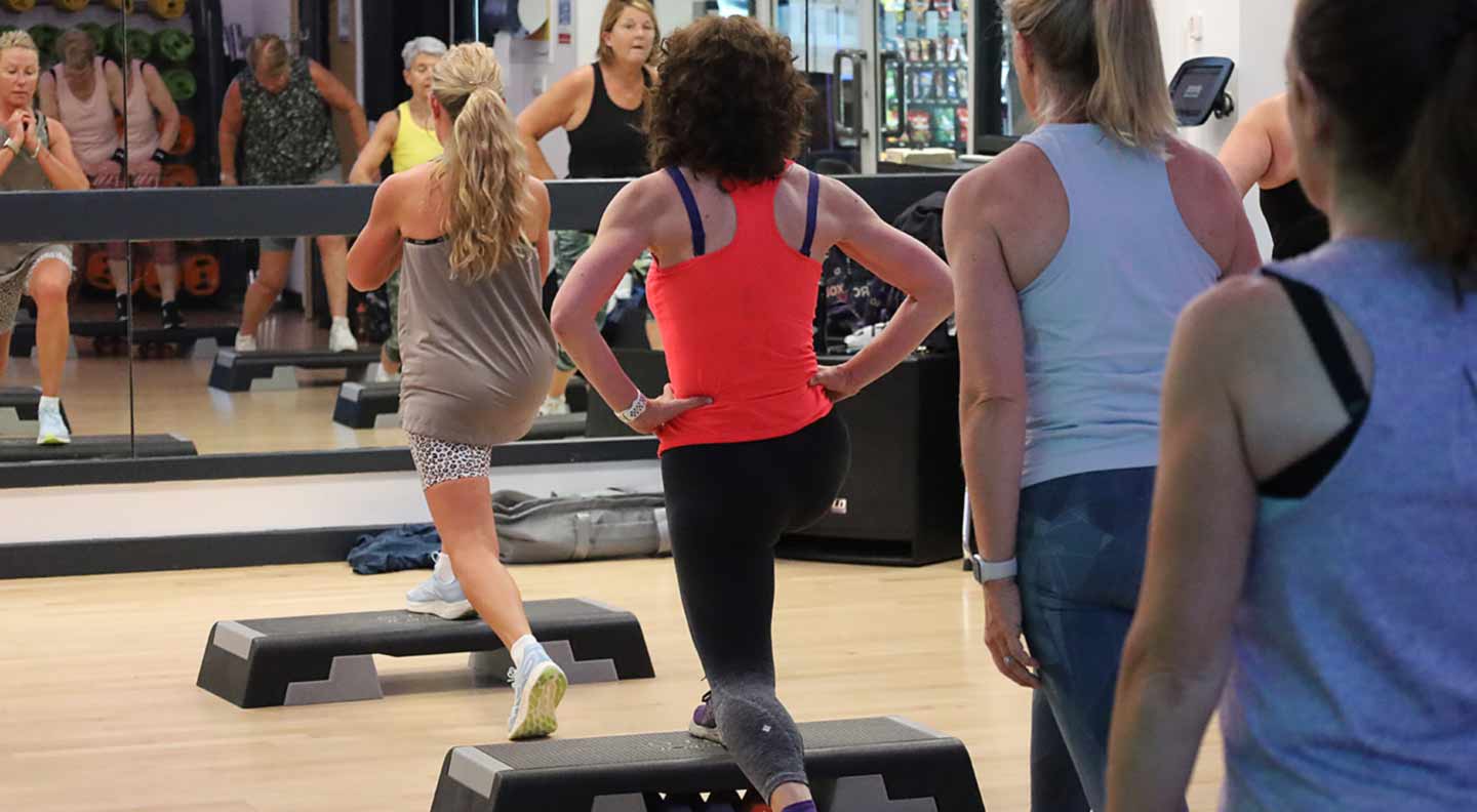 A group fitness class in a gym studio where several people are performing step‑up exercises on raised aerobic steps. Participants are positioned throughout the room facing a large wall mirror, and various gym equipment can be seen in the background.”