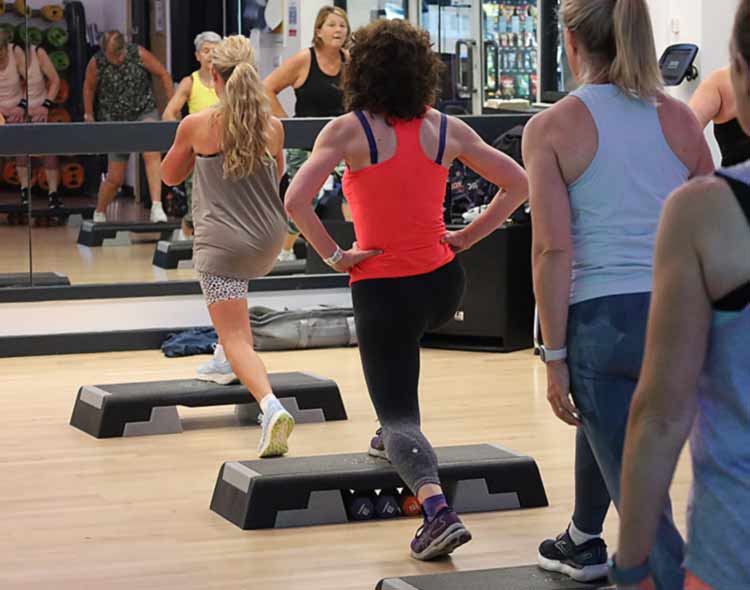 A group fitness class in a gym studio where several people are performing step‑up exercises on raised aerobic steps. Participants are positioned throughout the room facing a large wall mirror, and various gym equipment can be seen in the background.”