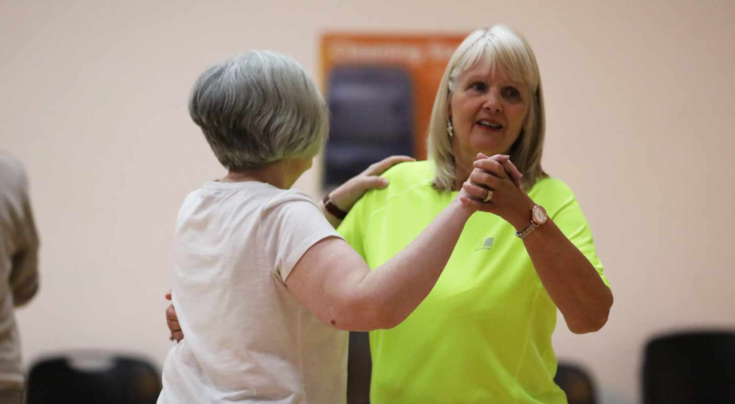 Two people are dancing together in a spacious indoor room. One person wearing a bright neon top holds the hands of another person in a light-colored shirt as they practice dance steps. Chairs and a wall poster are visible in the background, giving the setting the appearance of a community or fitness class.