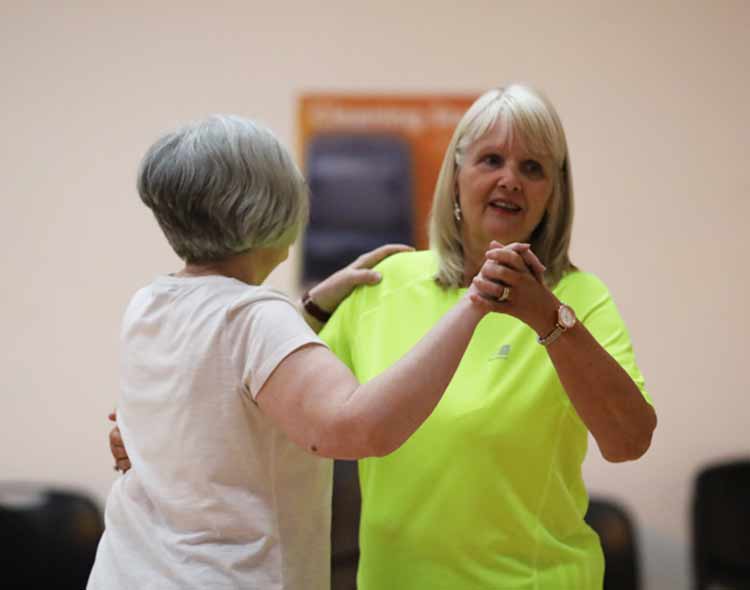 Two people are dancing together in a spacious indoor room. One person wearing a bright neon top holds the hands of another person in a light-colored shirt as they practice dance steps. Chairs and a wall poster are visible in the background, giving the setting the appearance of a community or fitness class.