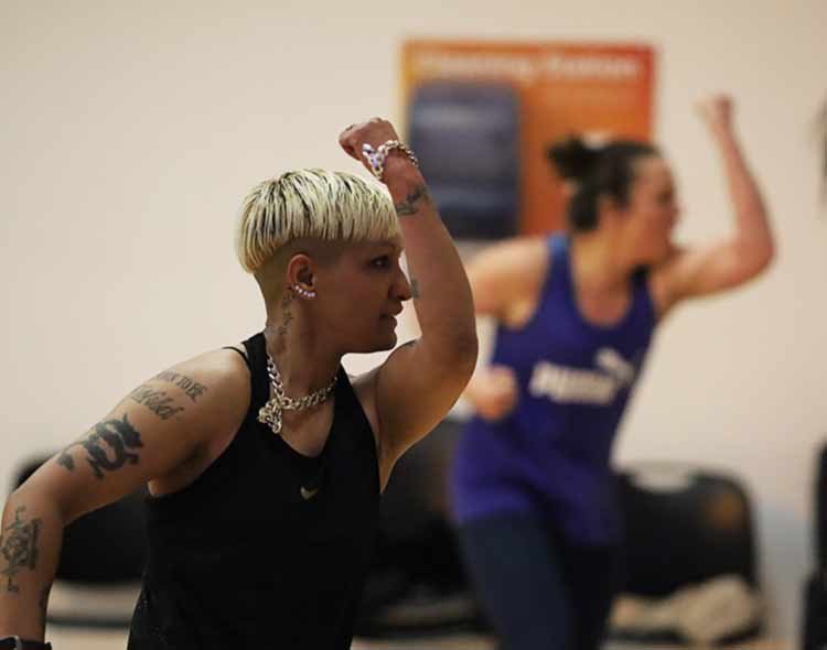 A group fitness class in motion, with a participant in the foreground raising an arm during an energetic routine.