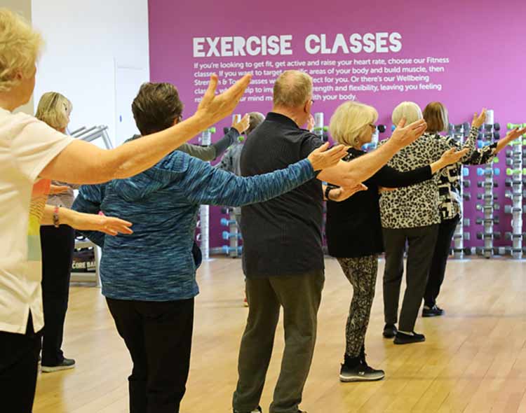 A group of participants in a studio following an exercise class routine with arms extended outward, facing a wall with large text that reads “EXERCISE CLASSES.”