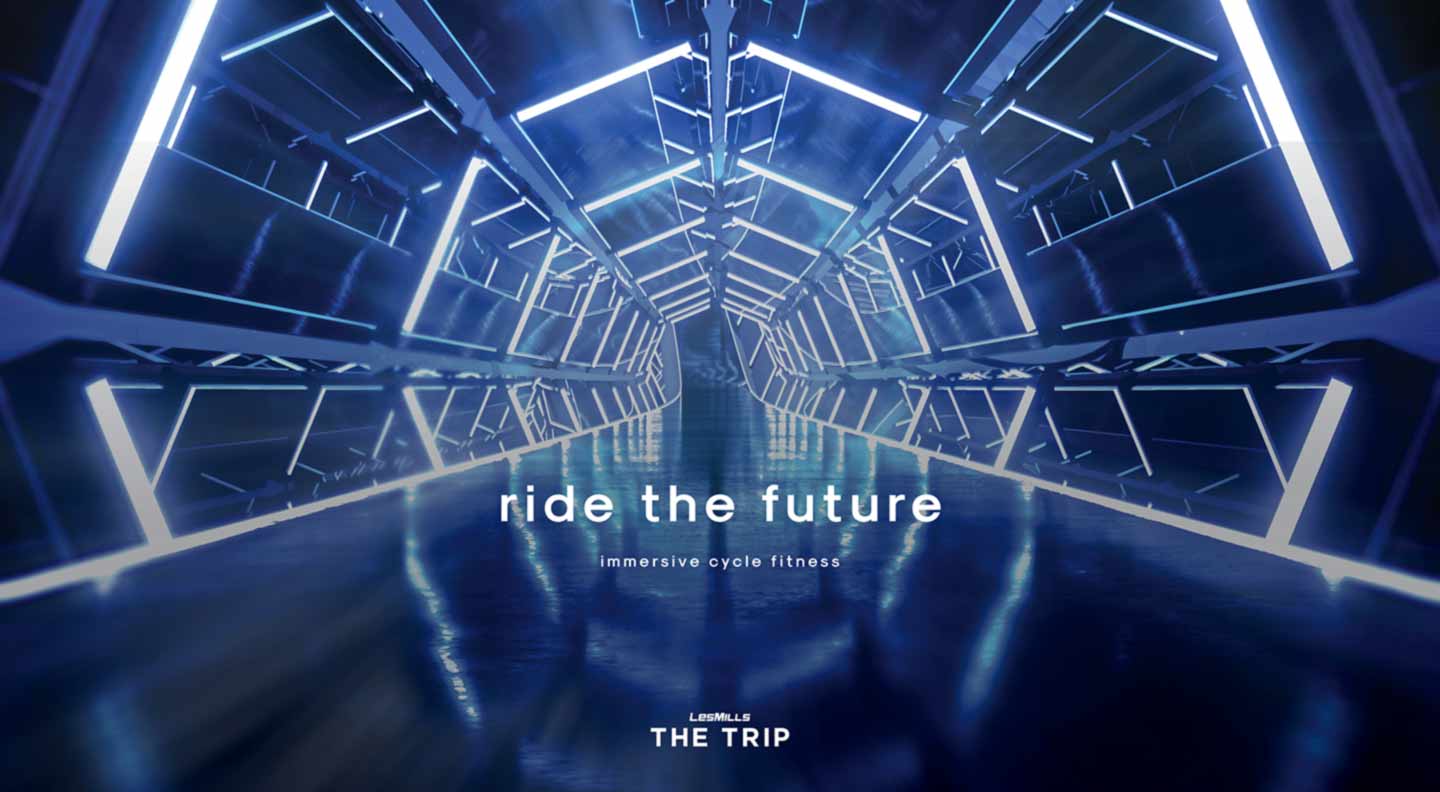 An illuminated futuristic tunnel with angular, metallic structures and bright blue-white LED light strips lines the walls and ceiling, creating a sense of depth and motion. Reflections on the glossy floor enhance the immersive, sci‑fi atmosphere. Centered text reads “ride the future” with the subtitle “immersive cycle fitness,” and below that the branding “LES MILLS THE TRIP.”