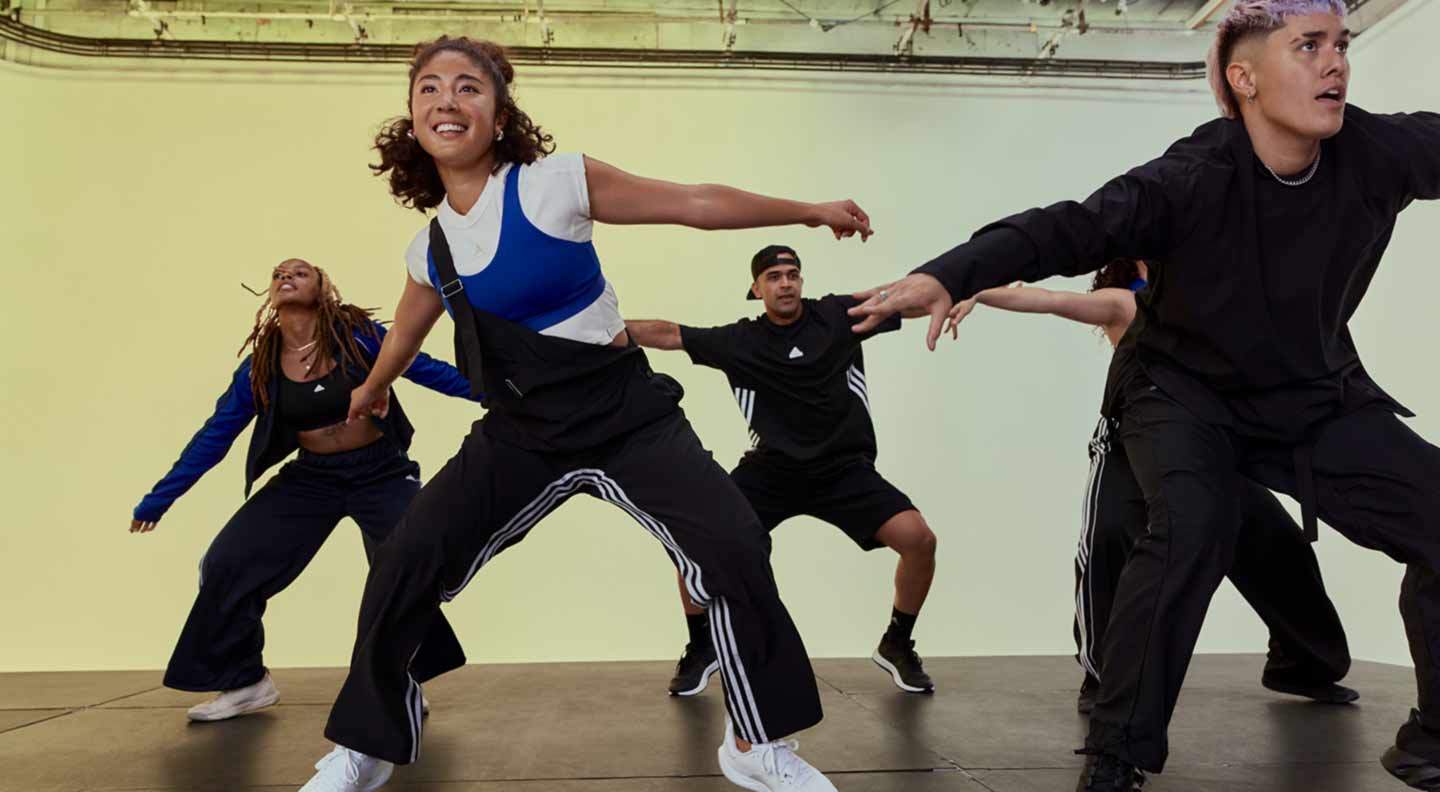 A group of people perform a dynamic dance routine on a studio stage with a smooth, dark floor. They are positioned in wide stances with arms extended outward in energetic, coordinated movements. The dancers wear athletic street‑style clothing, including loose‑fitting tops, track pants, and trainers. The background features a softly lit gradient wall and visible ceiling rigging with studio lights, giving the scene a professional, high‑energy rehearsal or class atmosphere.