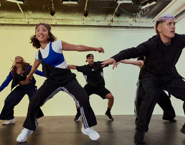 A group of people perform a dynamic dance routine on a studio stage with a smooth, dark floor. They are positioned in wide stances with arms extended outward in energetic, coordinated movements. The dancers wear athletic street‑style clothing, including loose‑fitting tops, track pants, and trainers. The background features a softly lit gradient wall and visible ceiling rigging with studio lights, giving the scene a professional, high‑energy rehearsal or class atmosphere.