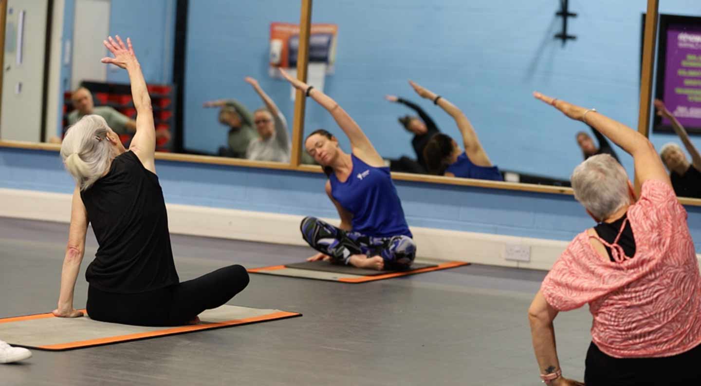 A group of people sit on exercise mats in a fitness studio, performing a seated side‑stretch with one arm reaching overhead. The room has light blue walls, a row of large mirrors reflecting the participants, and posters mounted on the wall. The floor is smooth and grey, and the participants are spaced evenly across the studio as they follow the stretching routine.