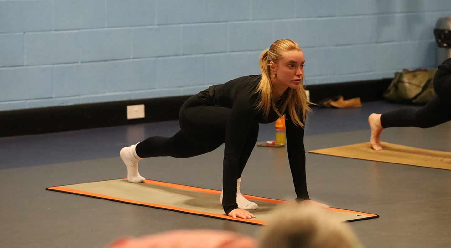 A person on a yoga mat performs a low lunge pose in an indoor fitness studio with light blue walls. Another participant on a nearby mat is in a similar position.