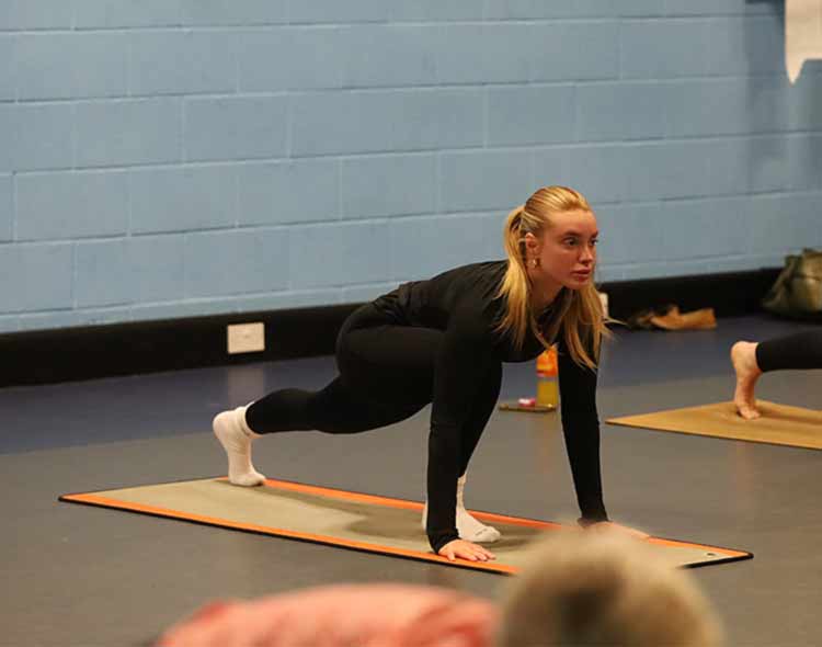A person on a yoga mat performs a low lunge pose in an indoor fitness studio with light blue walls. Another participant on a nearby mat is in a similar position.