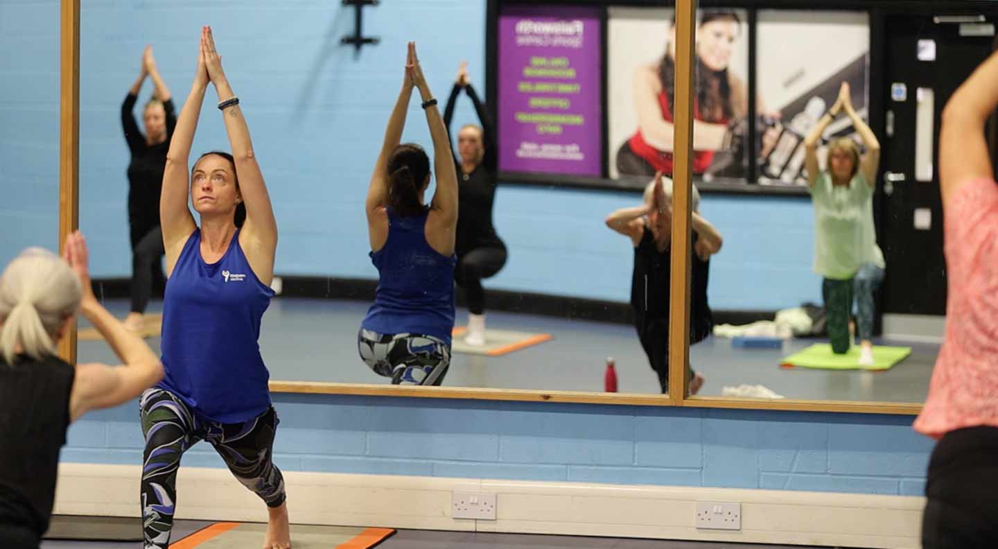 A group of people participate in a yoga or stretching class in a studio with light blue walls and large wall mirrors. The participants stand on exercise mats and hold a high lunge position with both arms raised overhead, forming a triangle shape with their hands. Reflections of the group are visible in the mirrors. Posters and equipment can be seen on the walls and in the background, and the scene is evenly lit with a clear view of the class formation.