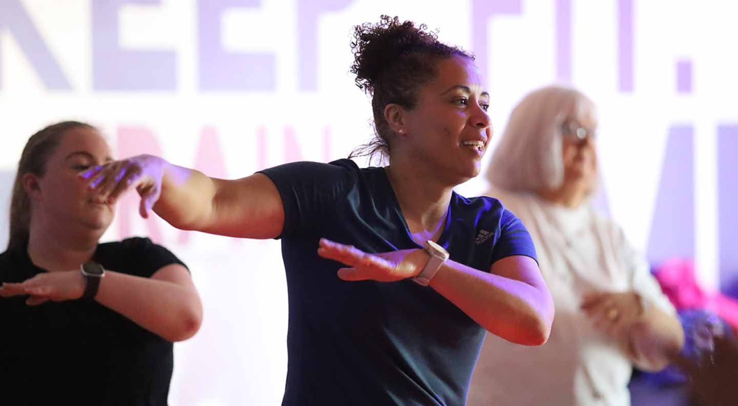 Several people participate in a group fitness class indoors. The person in the foreground extends both arms outward in a dynamic exercise pose, wearing a short-sleeved athletic top. Others in the background mirror similar movements. A large, brightly lit sign with bold lettering is partially visible behind them.