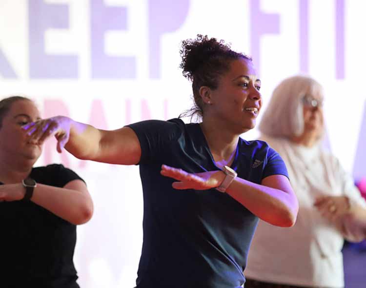 Several people participate in a group fitness class indoors. The person in the foreground extends both arms outward in a dynamic exercise pose, wearing a short-sleeved athletic top. Others in the background mirror similar movements. A large, brightly lit sign with bold lettering is partially visible behind them.