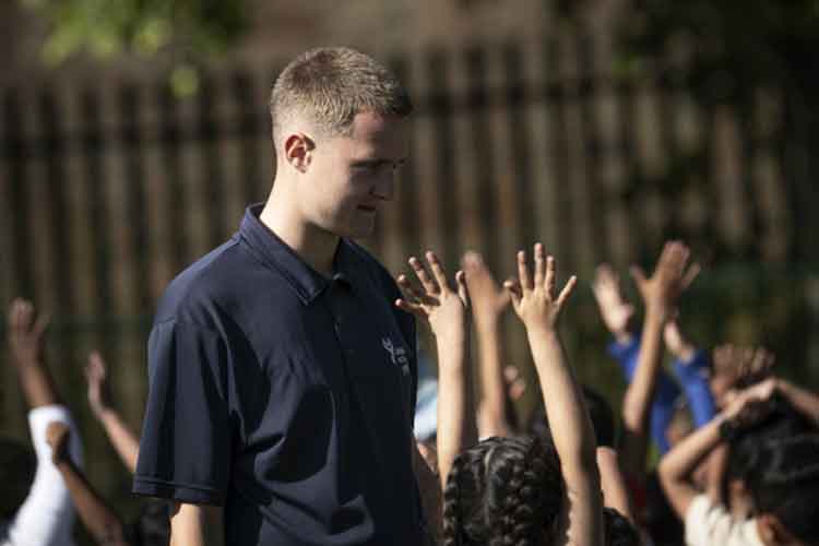 xMale instructor standing outdoors with a group of children raising their hands during an educational activity or school session.