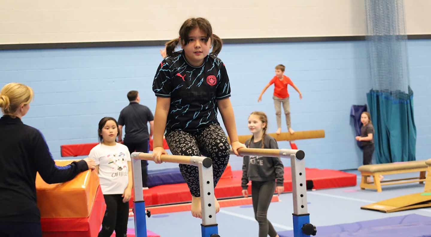 A young girl practices on parallel bars in a gymnasium while other children and instructors move around the room using various gymnastics equipment, including balance beams, mats, and soft play blocks