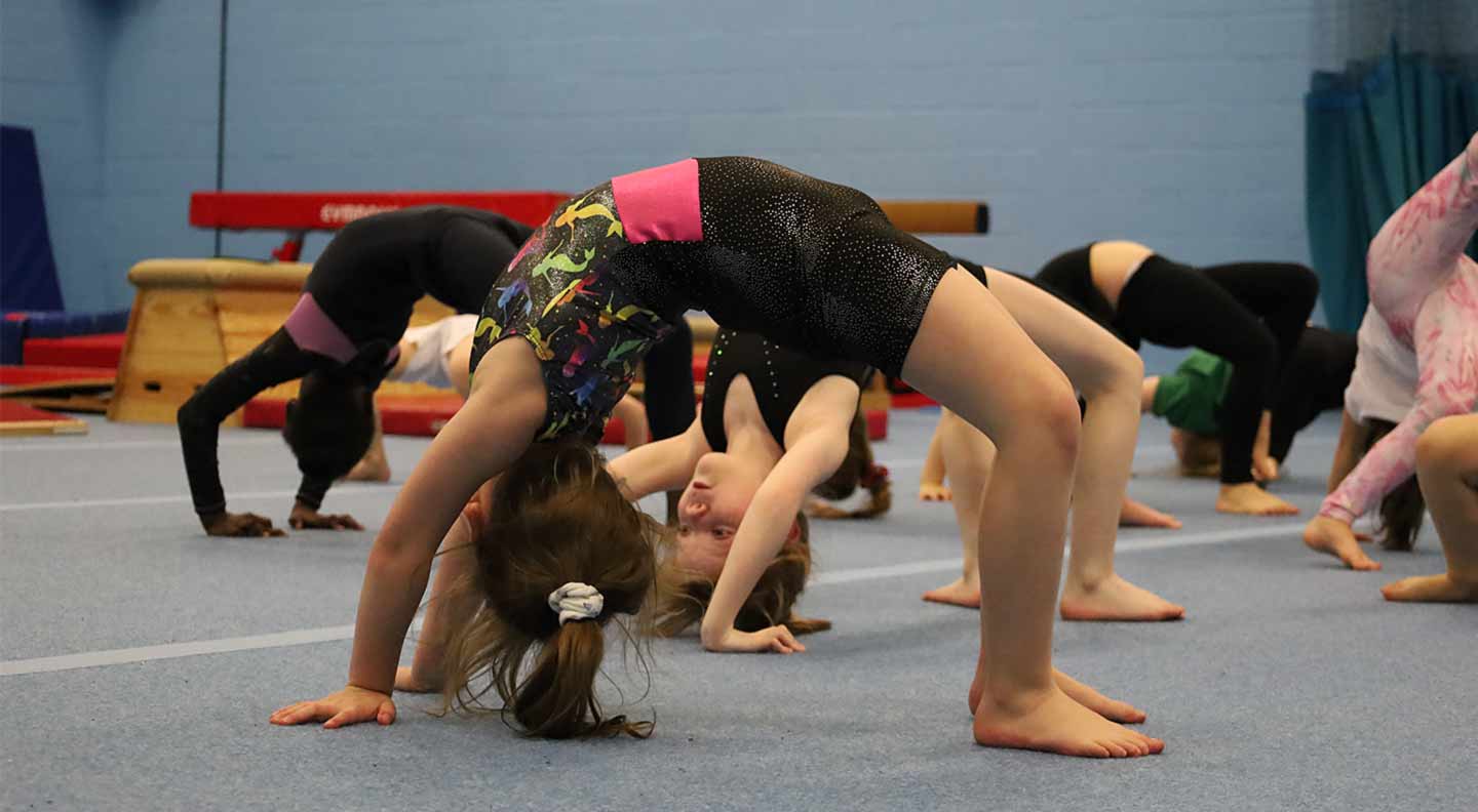 Children in a gymnastics class practise backbend bridge poses on a blue floor, with several gymnasts arched backward and supporting themselves on their hands and feet.