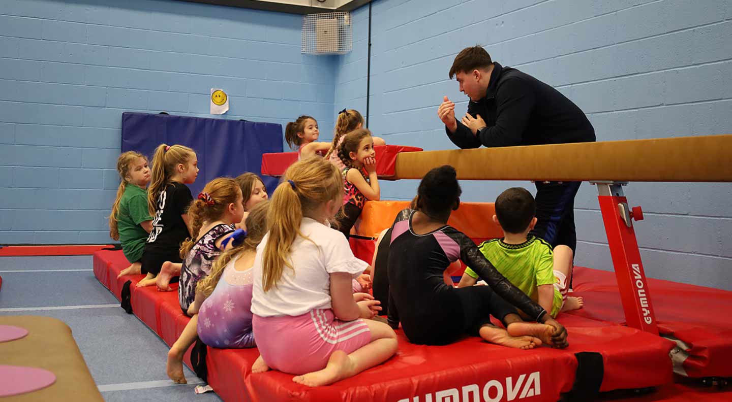 A coach speaks to a group of young gymnasts sitting on red mats around a balance beam, as they listen attentively during a training session in a sports hall.