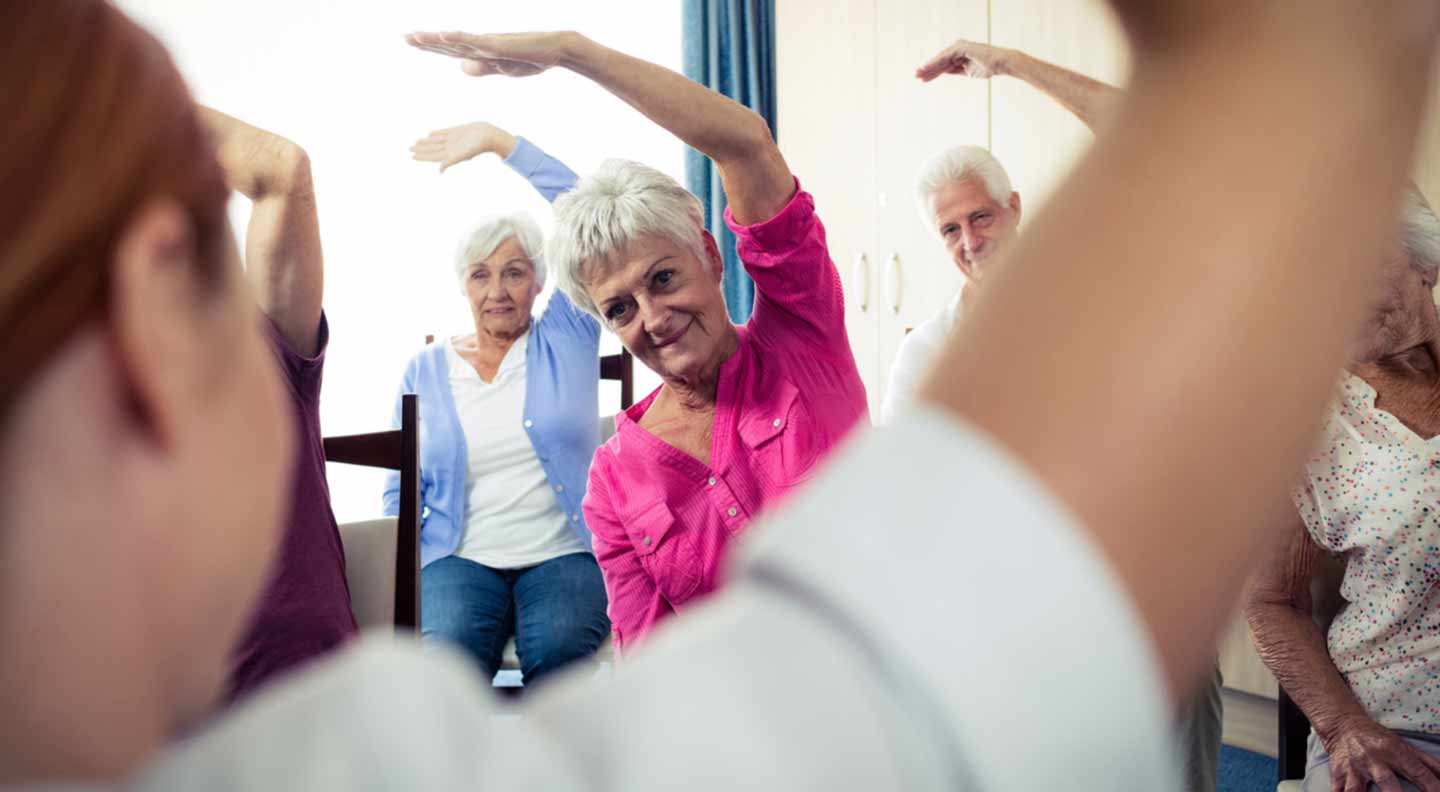 A group of older adults are seated in a circle during a gentle exercise or mobility class. They are raising one arm overhead and leaning to the side as part of a guided stretching routine. The instructor is positioned in the foreground with an arm lifted, mirroring the movement. The room appears to be a community or care setting, with chairs arranged for seated exercises and cabinets and curtains visible in the background.