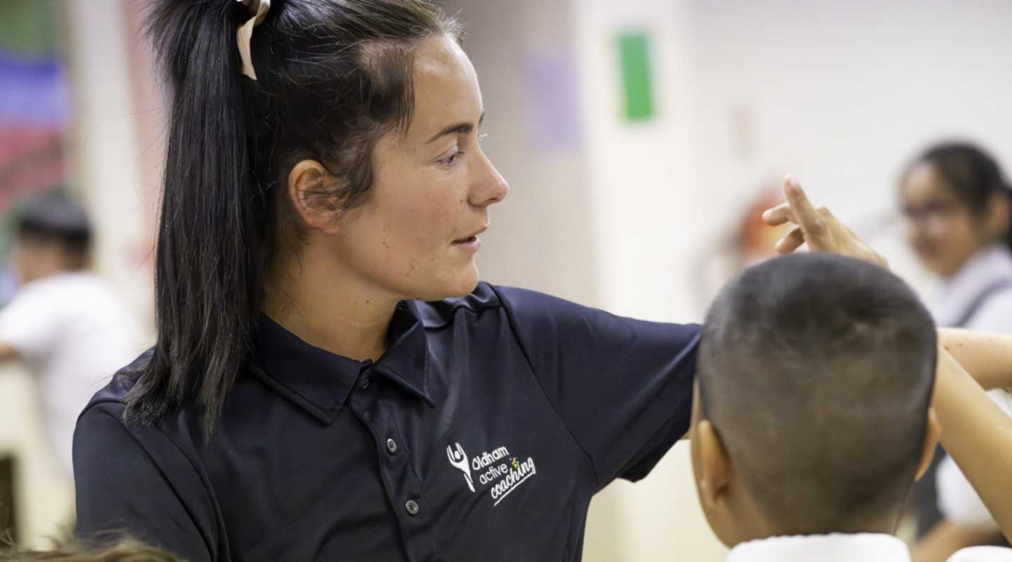 Oldham Active coach demonstrating a movement technique to a child during a school sports coaching session.