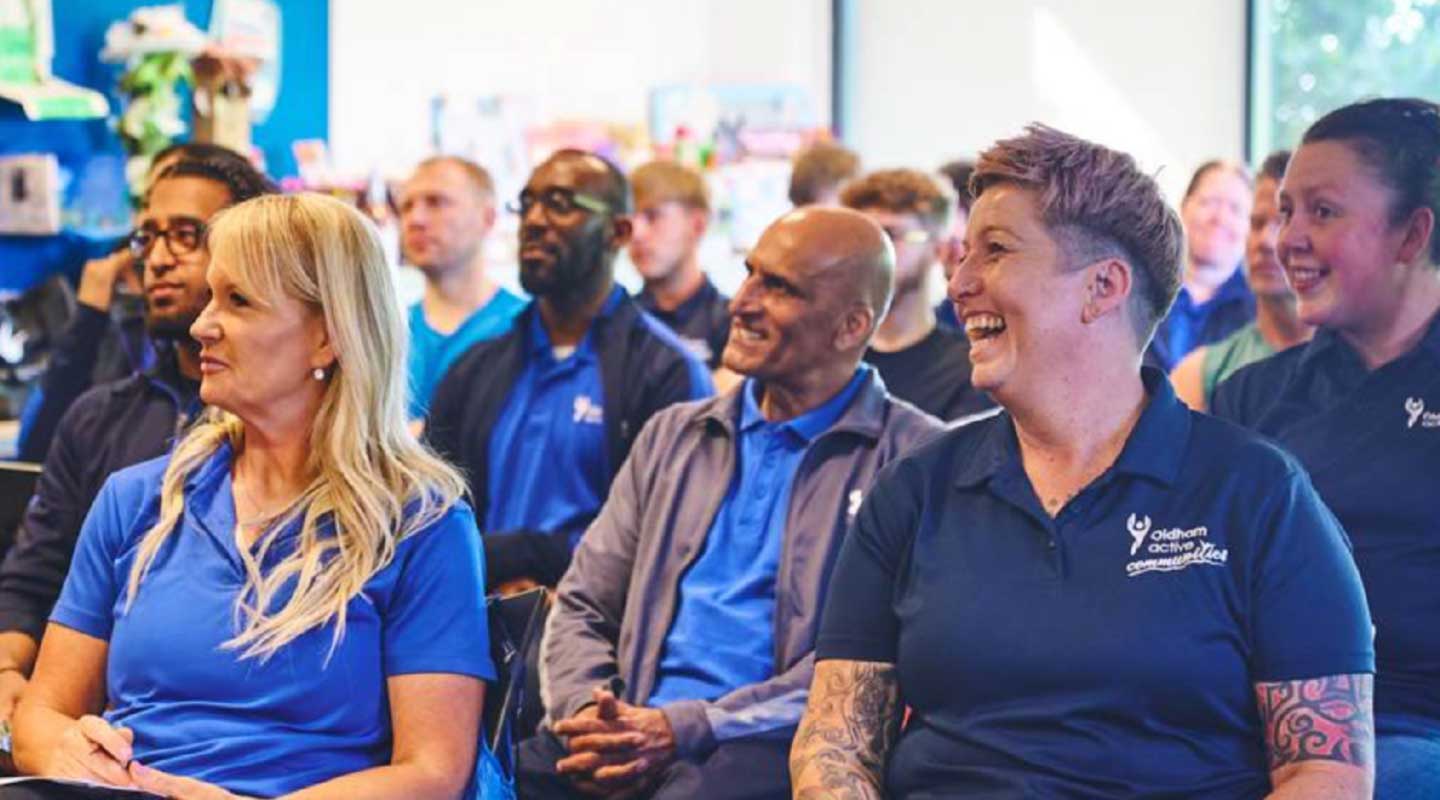 Group of adults wearing blue uniforms seated in a classroom-style training session, attentively listening and smiling during a team meeting or workshop.