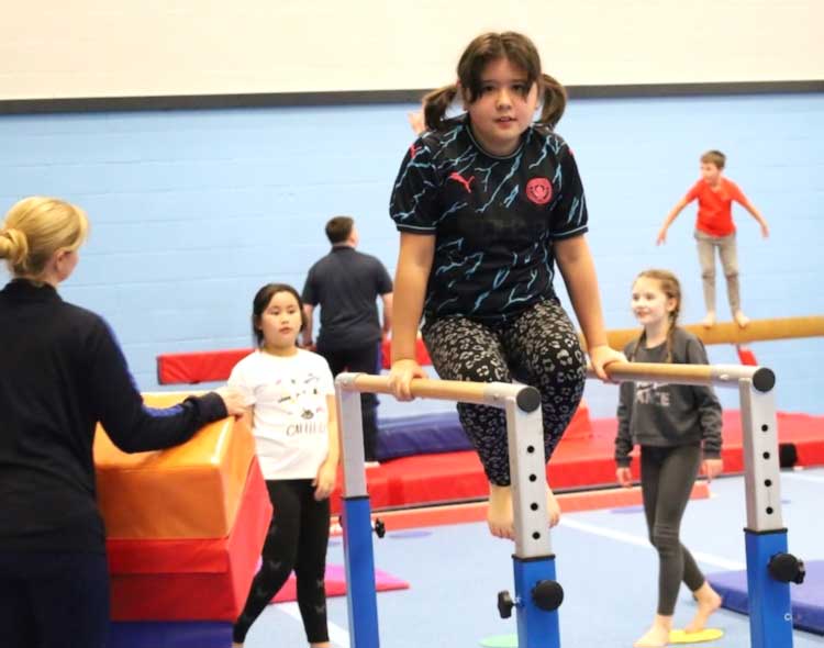 Girl on bar in gymnastics class, children in background trainign on mats